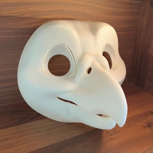 Könnte beinhalten: Eine weiße Vogelmaske mit langem Schnabel, Augenlöchern und einem lächelnden Mund. Die Maske besteht aus einem glatten Material und ist zum Tragen bestimmt. Die Maske liegt auf einer Holzoberfläche.