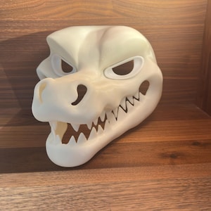 Puede incluir: Una máscara blanca con forma de dinosaurio, con dientes afilados y aberturas para los ojos. La máscara está hecha de un material sólido y probablemente sea para disfraz o cosplay. La máscara está diseñada para ser usada.