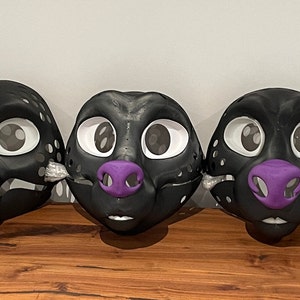 May include: Eight black and white fursuit heads with purple noses. The fursuit heads are arranged in a row on a wooden surface.