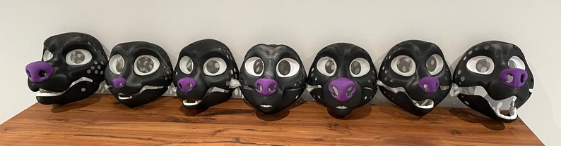 May include: Eight black and white fursuit heads with purple noses. The fursuit heads are arranged in a row on a wooden surface.