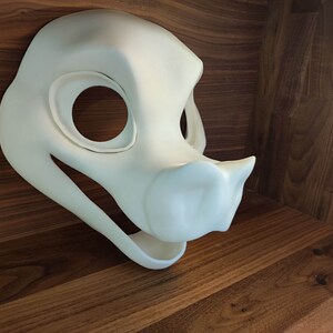 May include: A white, unpainted, furry animal mask with a large, rounded nose and open mouth. The mask is made of a smooth, hard material and has two large eye holes.