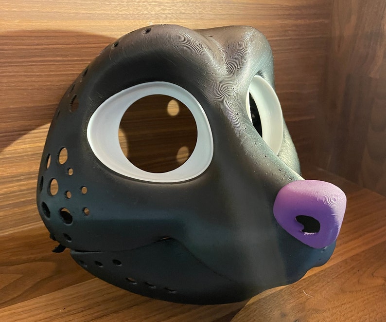 May include: A black furry animal mask with white eye holes and a purple nose. The mask is made of a smooth, textured material and has a detailed design.
