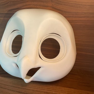 May include: White unpainted owl mask with large eye holes and a beak. The mask is made of a smooth, hard material.