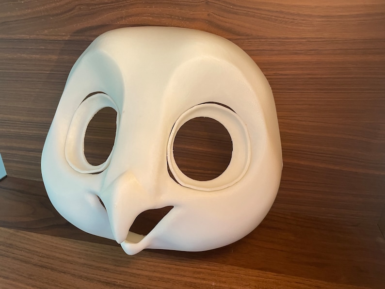 May include: White unpainted owl mask with large eye holes and a beak. The mask is made of a smooth, hard material.