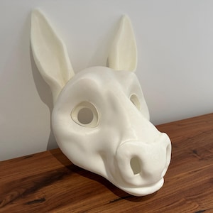 Könnte beinhalten: Eine weiße Eselmaske mit großen Ohren und Augenöffnungen. Die Maske besteht aus einem glatten Material und liegt auf einer Holzoberfläche. Die Maske ist eine Requisite für Kostüme oder Theateraufführungen.