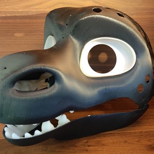 Könnte beinhalten: Eine schwarz-weiße 3D-gedruckte Maske, die einem Drachen- oder Dinosaurierkopf ähnelt. Die Maske hat große Augenöffnungen mit weißen Rahmen, eine detaillierte Schnauze und sichtbare Zähne. Sie hat mehrere kleine Löcher und eine glatte, strukturierte Oberfläche.