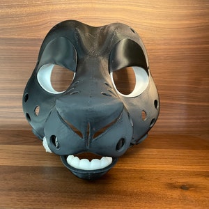 May include: A black and white furry animal mask with white eye holes and a black nose. The mask has a white mouth with teeth.