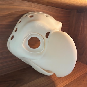 Könnte beinhalten: Eine cremefarbene Vogelmaske mit großem Schnabel und Augenöffnung. Die Maske hat mehrere kleine Löcher auf der Oberseite und an den Seiten. Die Maske besteht aus einem glatten Material, wahrscheinlich für Kostüm oder Cosplay.