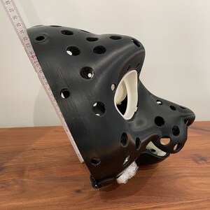 May include: A black 3D printed mask with a white interior. The mask has a series of holes throughout the surface. The mask is sitting on a wooden surface.