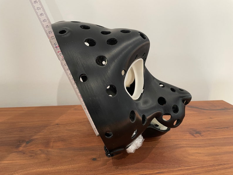 May include: A black 3D printed mask with a white interior. The mask has a series of holes throughout the surface. The mask is sitting on a wooden surface.