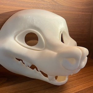 May include: White unpainted furry animal mask with a detailed snout and teeth. The mask has two eye holes and a large open mouth.