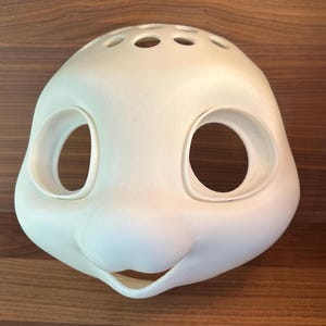 Könnte beinhalten: Weiße, unfertige Harzmaske mit zwei großen Augenlöchern und einem kleinen Mundloch. Die Maske hat eine abgerundete Form und mehrere kleine Löcher oben.