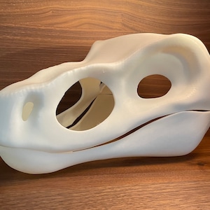 Könnte beinhalten: Eine weiße, 3D-gedruckte Dinosaurierkopfmaske mit offenen Augen- und Mundlöchern. Die Maske besteht aus glattem, weißem Kunststoff und hat ein realistisches Dinosaurierdesign.