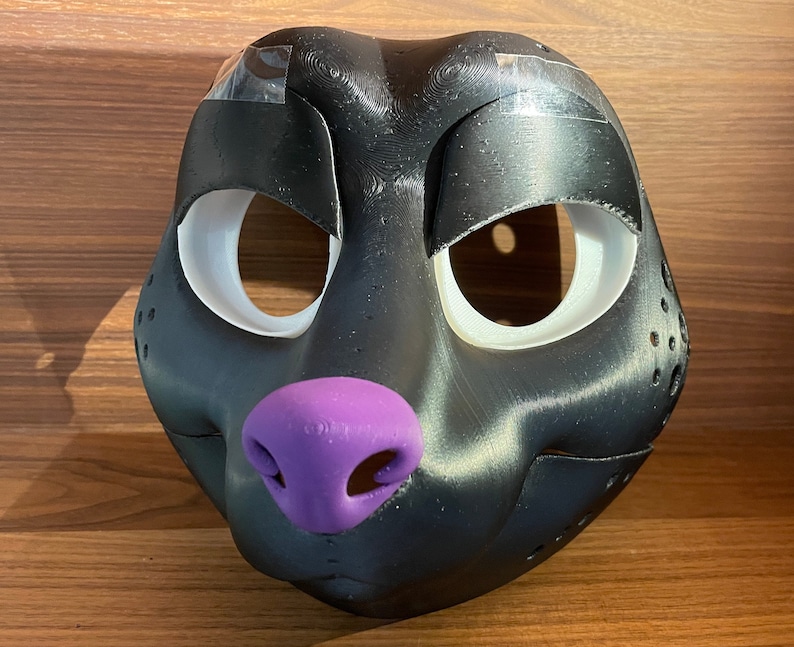 May include: A black furry animal mask with white eye holes and a purple nose. The mask is made of a smooth, textured material.