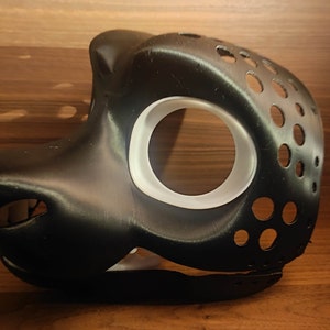 May include: A black and white furry mask with a large eye hole and a smaller nose hole. The mask is made of a smooth, hard plastic and has a textured surface.