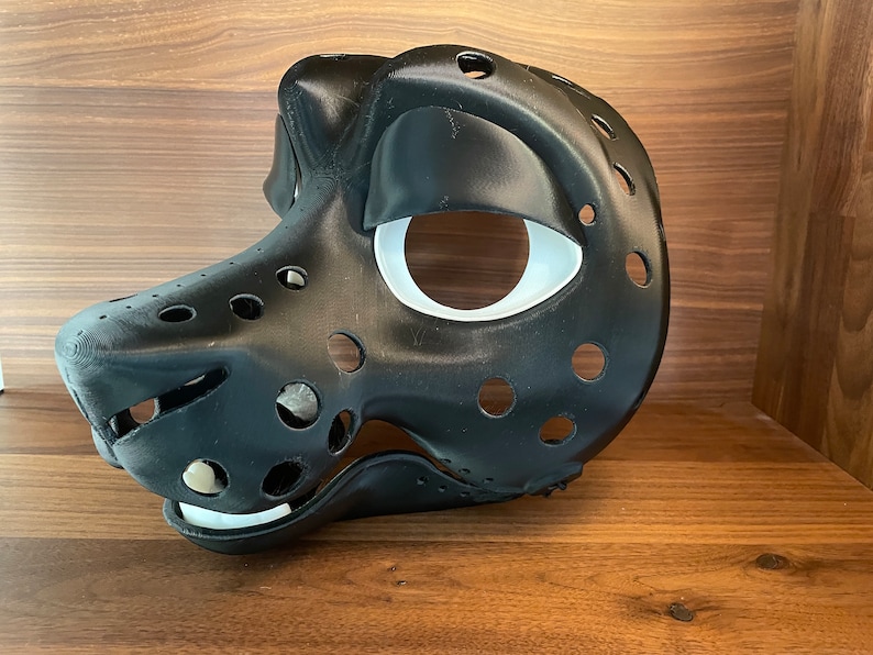 May include: A black and white furry animal mask with a white eye socket and a black nose. The mask is made of a hard plastic material and has a lot of holes in it.