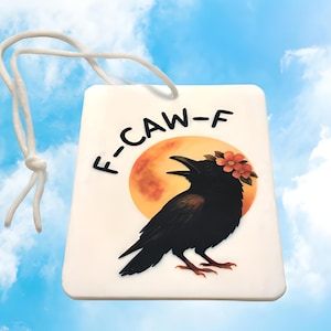 May include: A rectangular, off-white ornament with a black crow illustration, a full orange moon, and the text "F-CAW-F". The crow wears a floral crown. A white cord is attached for hanging. The background is a blue sky with clouds.
