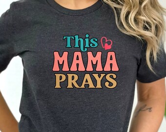 May include: A dark grey t-shirt with the text "This Mama Prays" in colorful retro lettering. The text includes a small red heart.