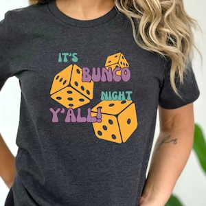 May include: A dark grey t-shirt with a graphic design of dice and the text "It's Bunco Night Y'all!"