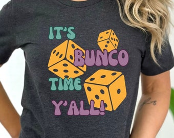 May include: A dark grey t-shirt with the text "It's Bunco Time Y'all!" in a retro font. The text is surrounded by four yellow dice with black dots.