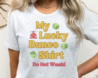 Könnte beinhalten: Ein graues T-Shirt mit dem Text "My Lucky Bunco Shirt" in Gelb und Rot. Auf dem Shirt sind grüne Würfel und ein rotes Glockensymbol abgebildet. Der Text "Do Not Wash!" ist in Rosa gedruckt.