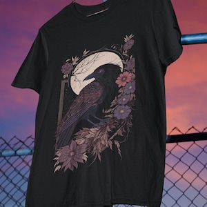 May include: Black t-shirt featuring a graphic of a raven perched on a branch with flowers and a crescent moon. The raven has red eyes and the flowers are purple and pink.