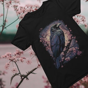 K&ouml;nnte beinhalten: Schwarzes T-Shirt mit einem grafischen Aufdruck eines Rabens, der auf einem Ast mit rosa Bl&uuml;ten sitzt. Der Rabe blickt nach links und hat eine dunkelblaue und schwarze Farbgebung.