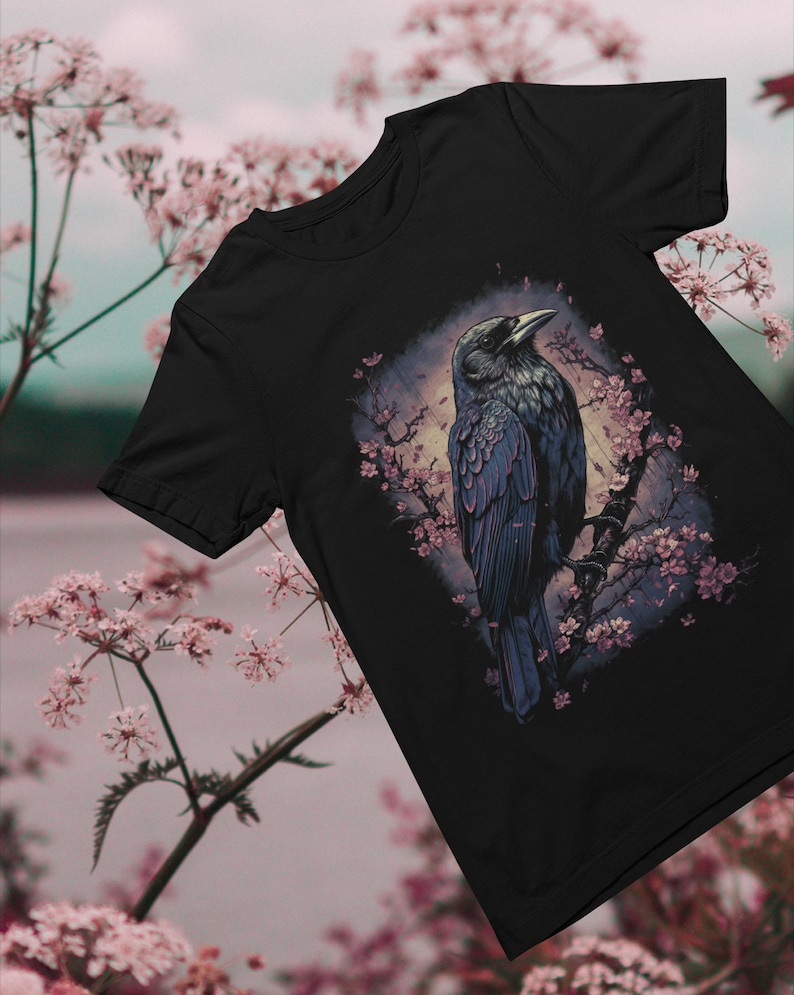 K&ouml;nnte beinhalten: Schwarzes T-Shirt mit einem grafischen Aufdruck eines Rabens, der auf einem Ast mit rosa Bl&uuml;ten sitzt. Der Rabe blickt nach links und hat eine dunkelblaue und schwarze Farbgebung.