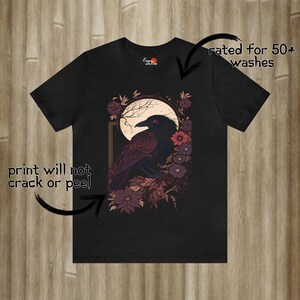 May include: Black t-shirt featuring a graphic print of a raven perched on a branch with flowers. The raven has red eyes and is surrounded by a moon and stars. The text "rated for 50+ washes" is on the right side of the image. The text "print will not crack or peel" is on the left side of the image.