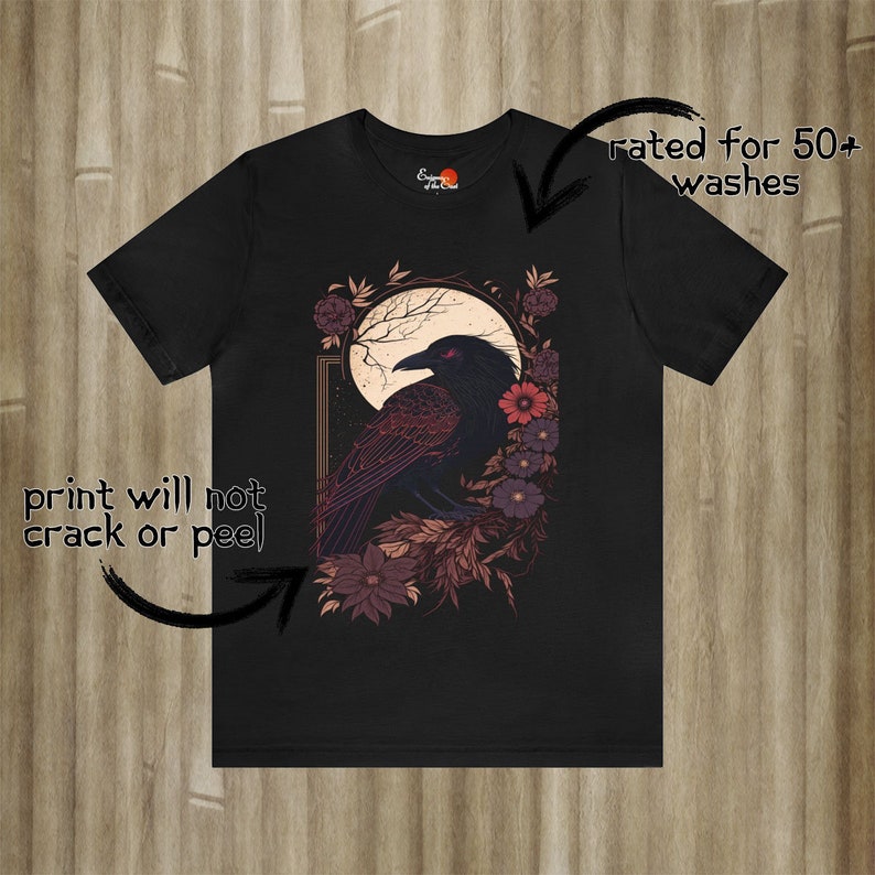 May include: Black t-shirt featuring a graphic print of a raven perched on a branch with flowers. The raven has red eyes and is surrounded by a moon and stars. The text "rated for 50+ washes" is on the right side of the image. The text "print will not crack or peel" is on the left side of the image.