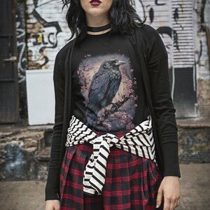 K&ouml;nnte beinhalten: Ein schwarzes T-Shirt mit einem Aufdruck eines Rabens, der auf einem Ast mit rosa Bl&uuml;ten sitzt. Das T-Shirt wird mit einem schwarzen Cardigan, einem gestreiften G&uuml;rtel und einem rot-schwarzen karierten Rock getragen.