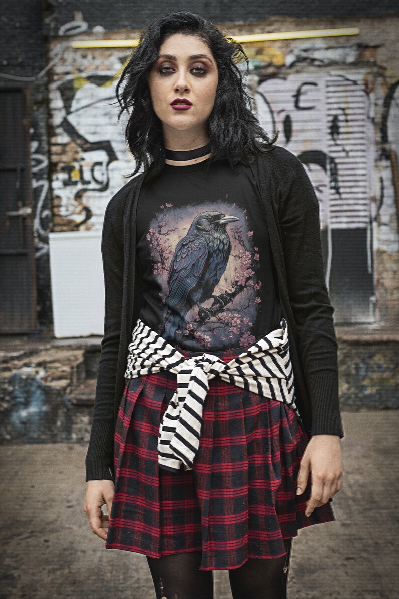 K&ouml;nnte beinhalten: Ein schwarzes T-Shirt mit einem Aufdruck eines Rabens, der auf einem Ast mit rosa Bl&uuml;ten sitzt. Das T-Shirt wird mit einem schwarzen Cardigan, einem gestreiften G&uuml;rtel und einem rot-schwarzen karierten Rock getragen.