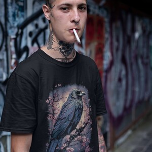 K&ouml;nnte beinhalten: Eine Person tr&auml;gt ein schwarzes T-Shirt mit einem grafischen Aufdruck eines Rabens, der auf einem Ast mit rosa Bl&uuml;ten sitzt. Der Rabe ist dunkelblau und grau mit einem schwarzen Schnabel. Die Person hat T&auml;towierungen an Hals und Gesicht.