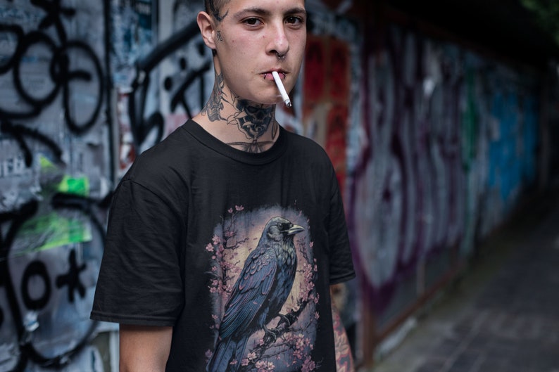 K&ouml;nnte beinhalten: Eine Person tr&auml;gt ein schwarzes T-Shirt mit einem grafischen Aufdruck eines Rabens, der auf einem Ast mit rosa Bl&uuml;ten sitzt. Der Rabe ist dunkelblau und grau mit einem schwarzen Schnabel. Die Person hat T&auml;towierungen an Hals und Gesicht.