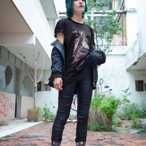 K&ouml;nnte beinhalten: Schwarzes T-Shirt mit Grafikdesign, kombiniert mit schwarzen Leggings und Schuhen. Eine dunkelblaue Jacke wird &uuml;ber dem Shirt getragen. Die Person hat gr&uuml;ne Haare und steht auf einer roten Ziegeloberfl&auml;che.