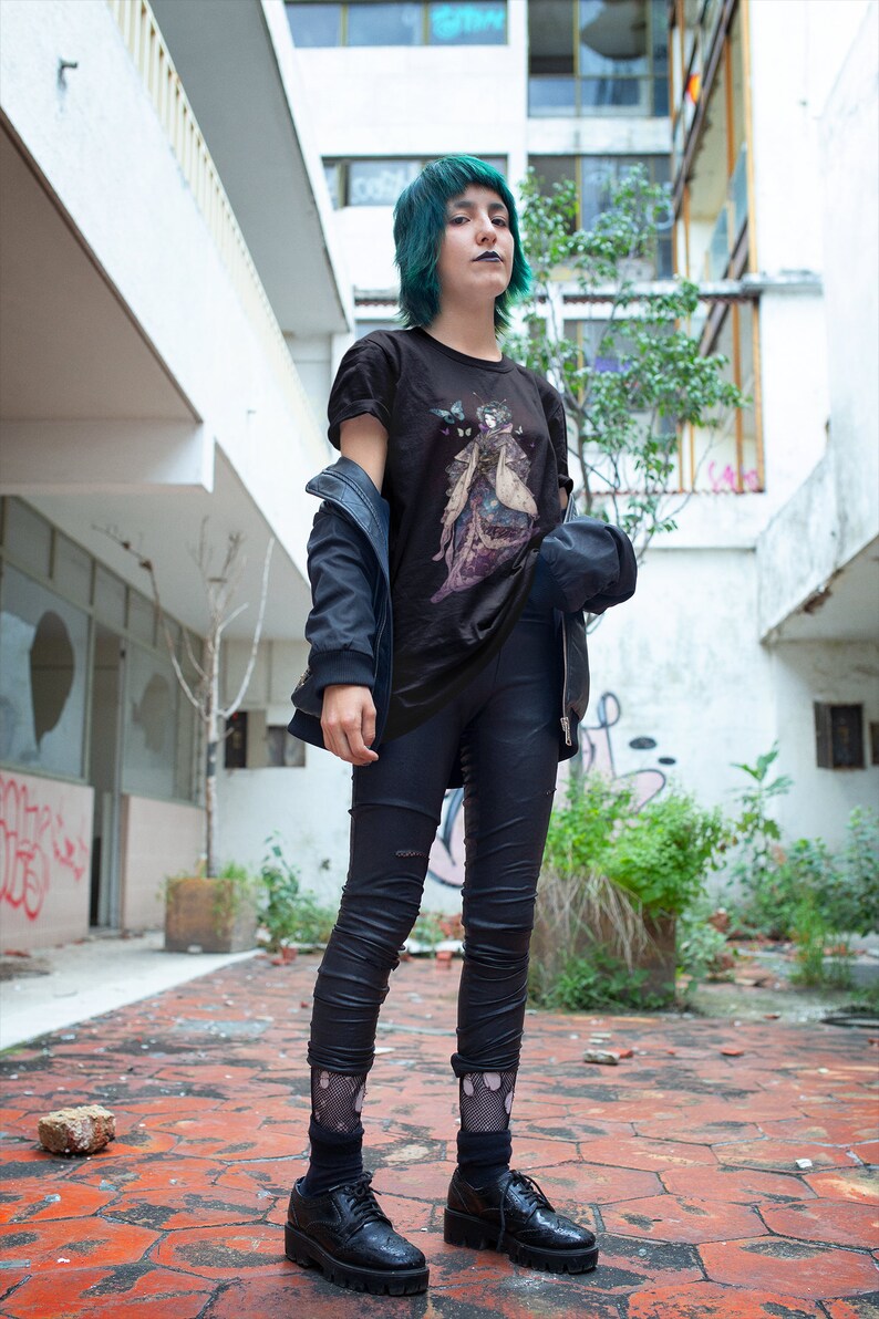 K&ouml;nnte beinhalten: Schwarzes T-Shirt mit Grafikdesign, kombiniert mit schwarzen Leggings und Schuhen. Eine dunkelblaue Jacke wird &uuml;ber dem Shirt getragen. Die Person hat gr&uuml;ne Haare und steht auf einer roten Ziegeloberfl&auml;che.