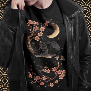 May include: Black cat lounging on a branch of a cherry blossom tree with a crescent moon in the background. The cat is surrounded by pink blossoms and stars. The design is on a black t-shirt.