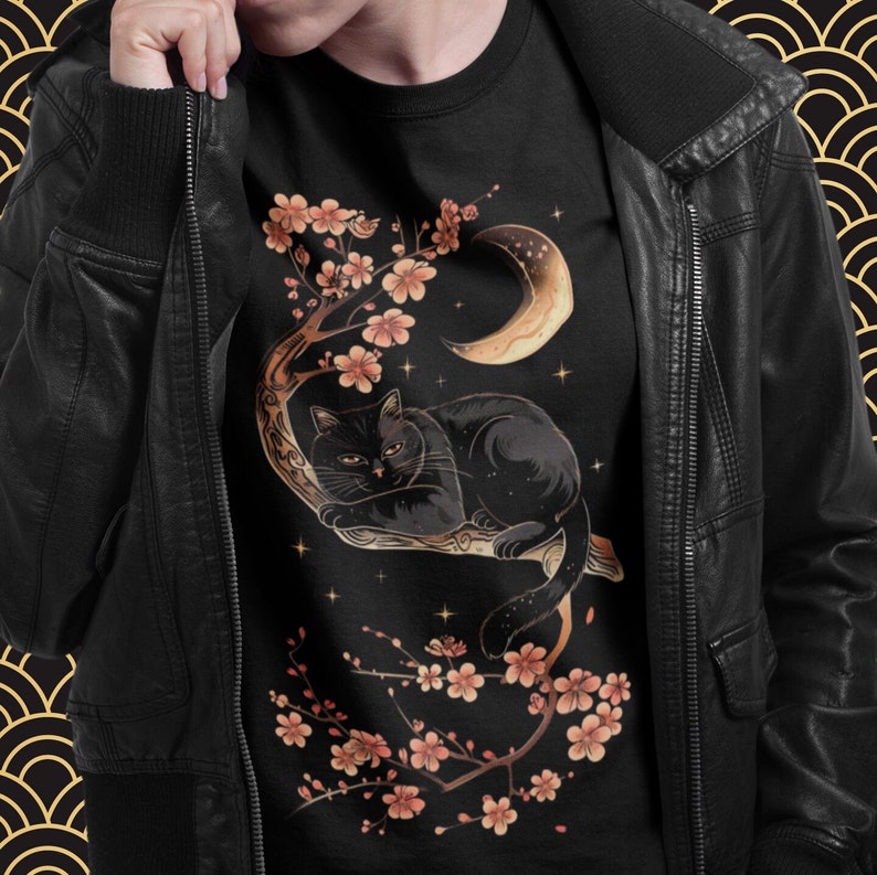 May include: Black cat lounging on a branch of a cherry blossom tree with a crescent moon in the background. The cat is surrounded by pink blossoms and stars. The design is on a black t-shirt.