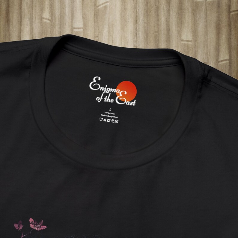 K&ouml;nnte beinhalten: Schwarzes T-Shirt mit einem wei&szlig;en Etikett, auf dem "Enigma of the East" mit einem roten Kreis steht. Das Etikett enth&auml;lt auch die Gr&ouml;&szlig;e "L" und den Text "100% Baumwolle Hergestellt in Bangladesch".