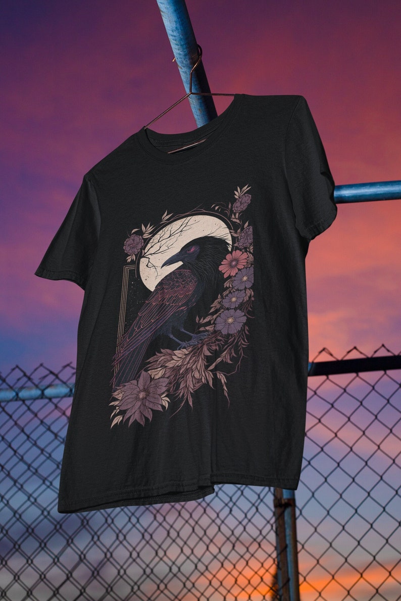 May include: Black t-shirt featuring a graphic of a raven perched on a branch with flowers and a crescent moon. The raven has red eyes and the flowers are purple and pink.