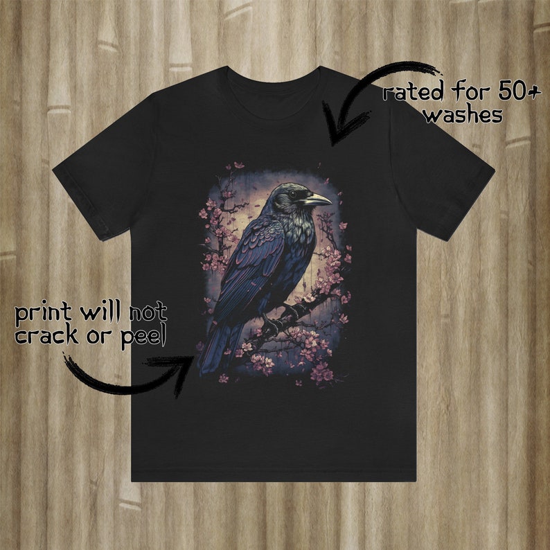 K&ouml;nnte beinhalten: Schwarzes T-Shirt mit einem grafischen Aufdruck eines Rabens, der auf einem Ast mit rosa Bl&uuml;ten sitzt. Der Druck ist f&uuml;r 50+ W&auml;schen geeignet und wird nicht rei&szlig;en oder abbl&auml;ttern.