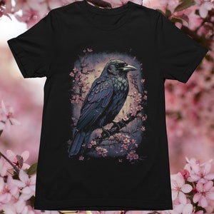 K&ouml;nnte beinhalten: Ein schwarzer Rabe sitzt auf einem Ast mit rosa Bl&uuml;ten. Der Rabe blickt nach rechts und hat einen dunkelblaugrauen K&ouml;rper mit einem schwarzen Schnabel. Der Hintergrund ist ein verschwommenes Bild eines Waldes.