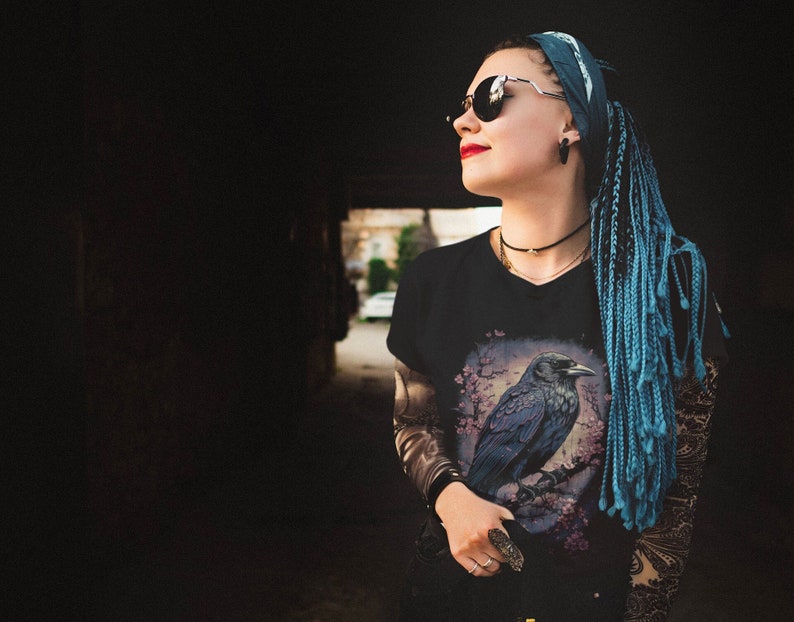K&ouml;nnte beinhalten: Eine Frau mit blauen Z&ouml;pfen und einem schwarzen Bandana tr&auml;gt ein schwarzes T-Shirt mit einem Motiv eines Rabens auf einem Ast. Sie tr&auml;gt eine Sonnenbrille und einen Ring an ihrem Finger.
