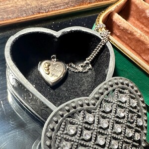 May include: A heart-shaped metal trinket box with a textured, rhinestone-encrusted lid. Inside, a silver-toned heart locket hangs on a delicate chain. The locket features three small gold-colored hearts and the engraved text I Love You.