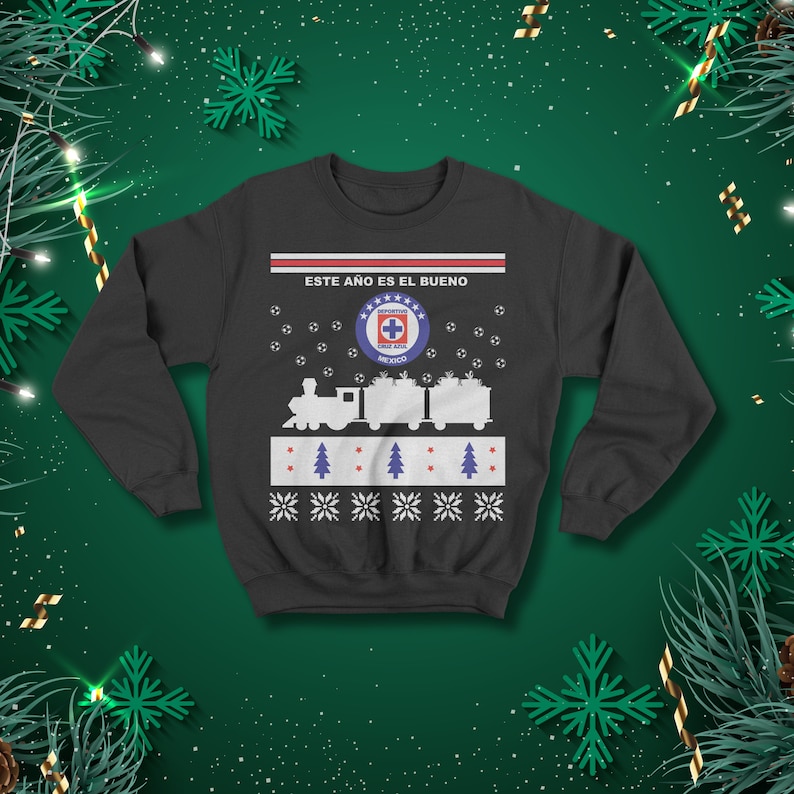 May include: A dark grey sweatshirt with a festive design. The design includes a train, footballs, and the text "ESTE A&Ntilde;O ES EL BUENO". The sweatshirt also features the Cruz Azul Mexico logo and snowflake patterns.