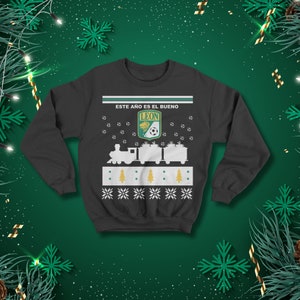 May include: Dark grey sweatshirt with a festive design. The design includes a train carrying gifts, soccer balls, and the text "ESTE A&Ntilde;O ES EL BUENO" above the team logo. The sweatshirt has a green and white colour scheme.