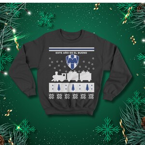 May include: Dark grey sweatshirt featuring a festive design. The design includes a train carrying gifts, footballs, Christmas trees, and snowflakes. The text "ESTE A&Ntilde;O ES EL BUENO" is printed across the top.