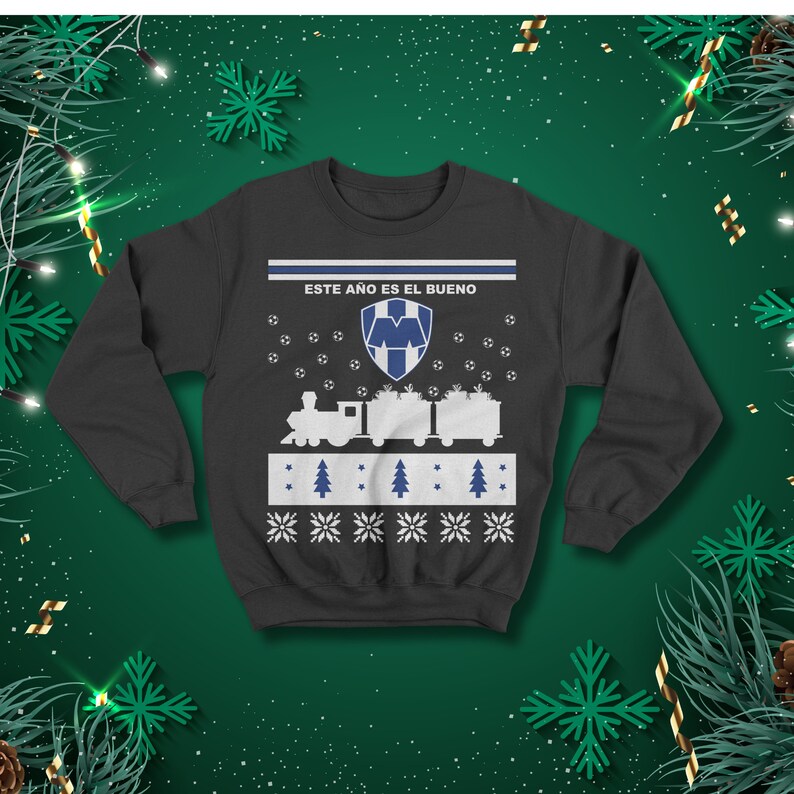 May include: Dark grey sweatshirt featuring a festive design. The design includes a train carrying gifts, footballs, Christmas trees, and snowflakes. The text "ESTE A&Ntilde;O ES EL BUENO" is printed across the top.