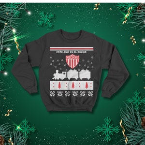 May include: Dark grey sweatshirt with a festive design. The design includes a train, soccer balls, snowflakes, and the text "ESTE A&Ntilde;O ES EL BUENO" above a team logo. The sweatshirt has a crew neck and long sleeves.