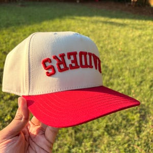 May include: A cream-colored baseball cap with a flat red brim. The front panel features the word RAIDERS embroidered in bold, red, capital letters, positioned upside down. The cap has a structured crown and a stitched red brim.